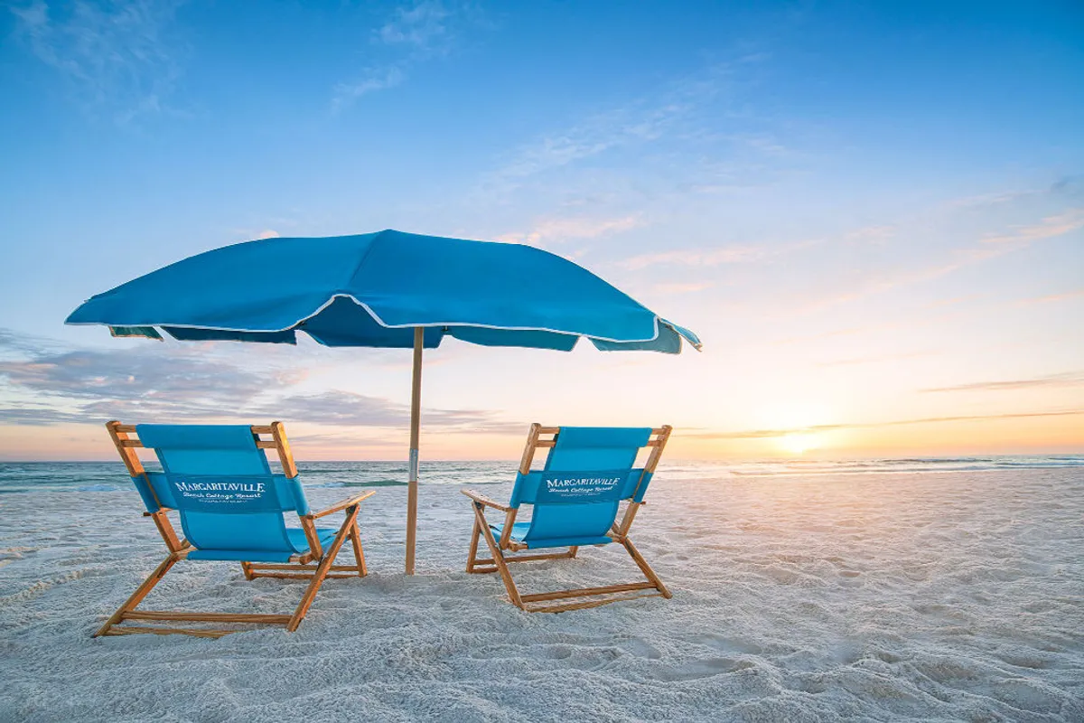 The image showcases two iconic blue Margaritaville-branded beach chairs and a matching umbrella set on soft, white sand, facing a tranquil ocean as the sun sets over the horizon. The scene evokes a serene, inviting atmosphere perfect for relaxation and beachfront leisure.