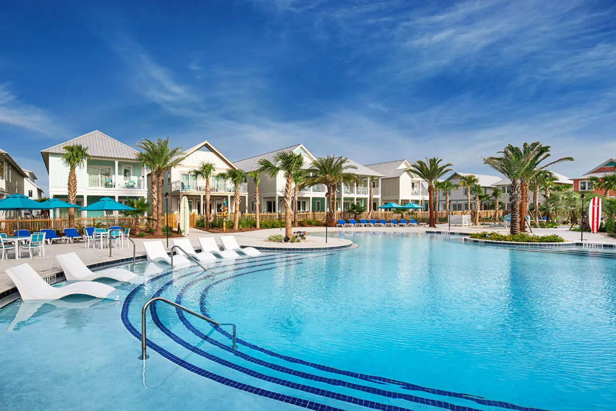 A large resort-style swimming pool with semi-submerged lounge chairs, two-story beach cottage villas with balconies in the background, palm trees, and poolside umbrellas. The area is filled with natural light and exudes a relaxed vacation ambiance.