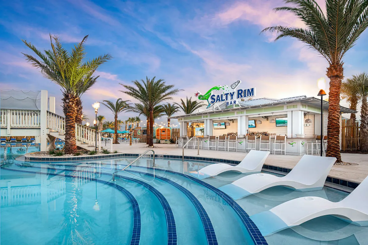 This image showcases the Salty Rim Bar with its inviting open-air design, the curved swimming pool featuring built-in lounge chairs, and tall palm trees swaying above. The lively pool area is accented by a staircase and comfortable seating arrangements, setting the scene for socializing and relaxation.