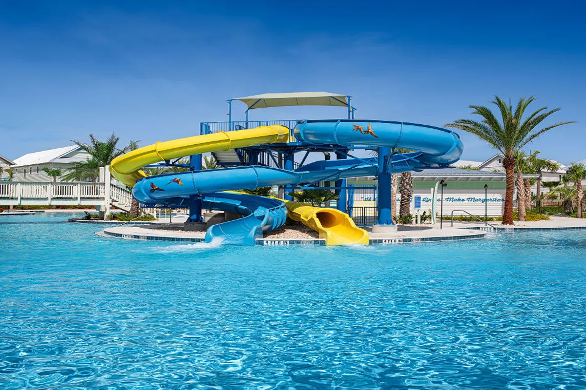 A vibrant pool scene centered on colorful yellow and blue water slides, a large circular pool, palm trees, and resort buildings in the background. Guests can be seen swimming, sliding, and enjoying the lively atmosphere.