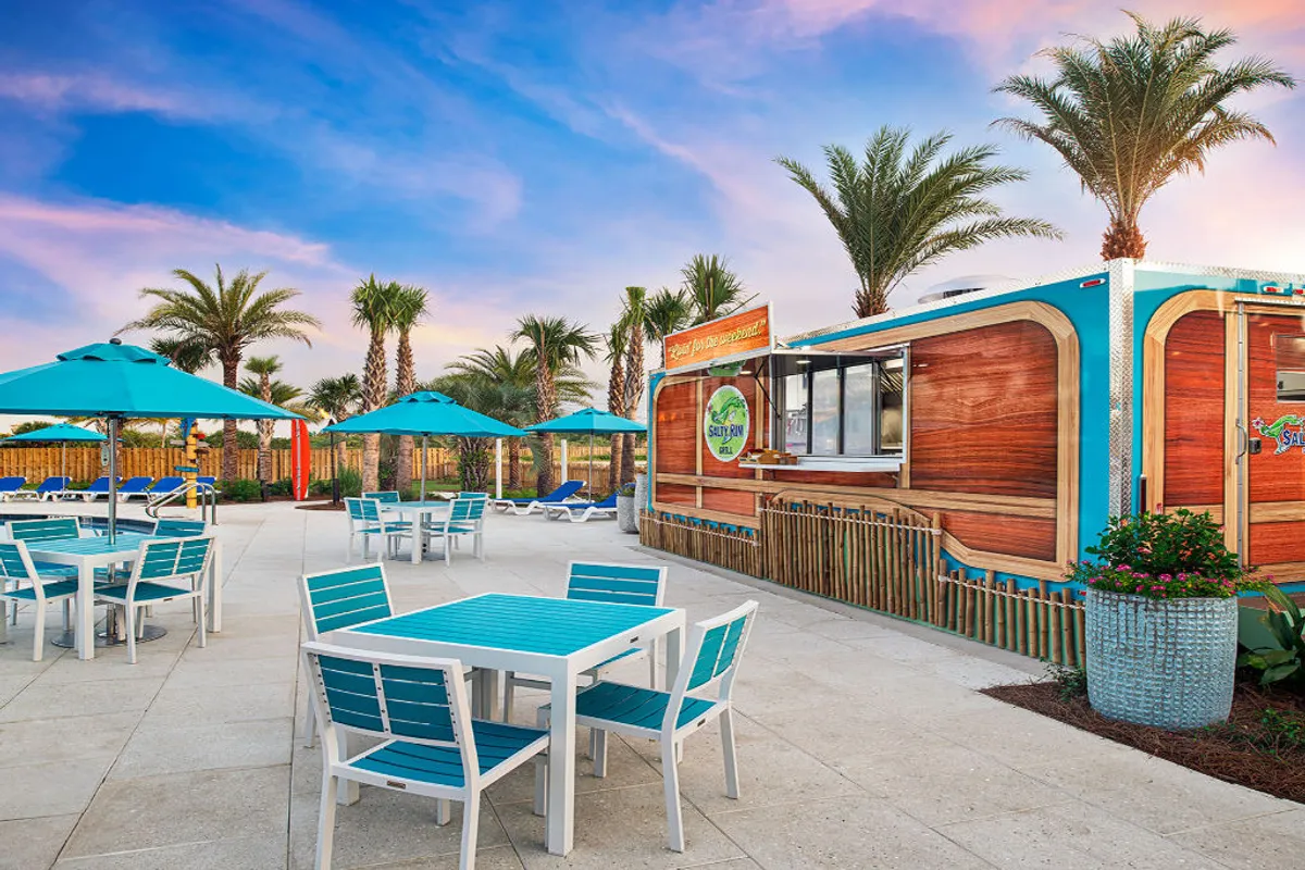 A bright, tropical-themed poolside dining area with turquoise patio tables and umbrellas sits adjacent to a wood-paneled concession stand or bar. Palm trees and lounge chairs dot the background, inviting guests to relax, dine, or enjoy refreshments in a casual outdoor setting.