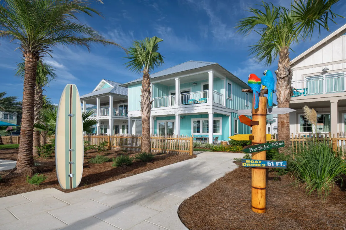 This image showcases a turquoise two-story beach villa with balconies, framed by palm trees and playful tropical decor, including a colorful wooden signpost with parrots and a large upright surfboard. The villa’s walkways and balconies hint at private relaxation spaces with direct access to the resort’s outdoor amenities.