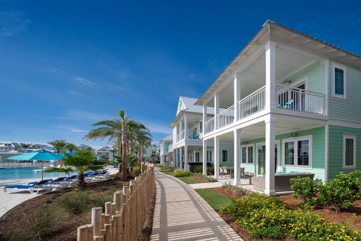 Pastel-colored beachfront cottages with inviting balconies overlook private pool areas complete with blue umbrellas, lounge chairs, and lush landscaping. The scene shows guests sunbathing, swimming, and relaxing poolside, just steps from the beach pathway.