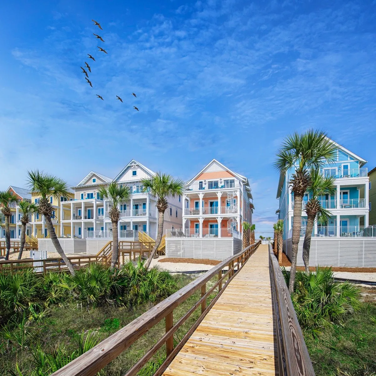 Colorful three-story beach houses with balconies are lined up, with a wooden boardwalk leading towards the beach. Palm trees frame the houses, and a flock of birds can be seen flying overhead, evoking a lively and carefree beach holiday atmosphere.