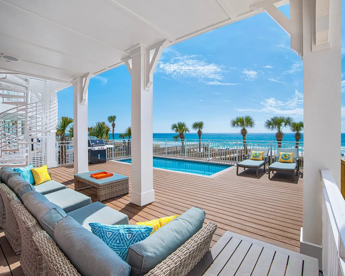 A vibrant outdoor pool deck scene with a private heated swimming pool, palm trees, ocean views, and a comfortable outdoor sectional sofa with colorful cushions. The spiral staircase and sun-drenched patio create an inviting space for both relaxation and social gatherings.