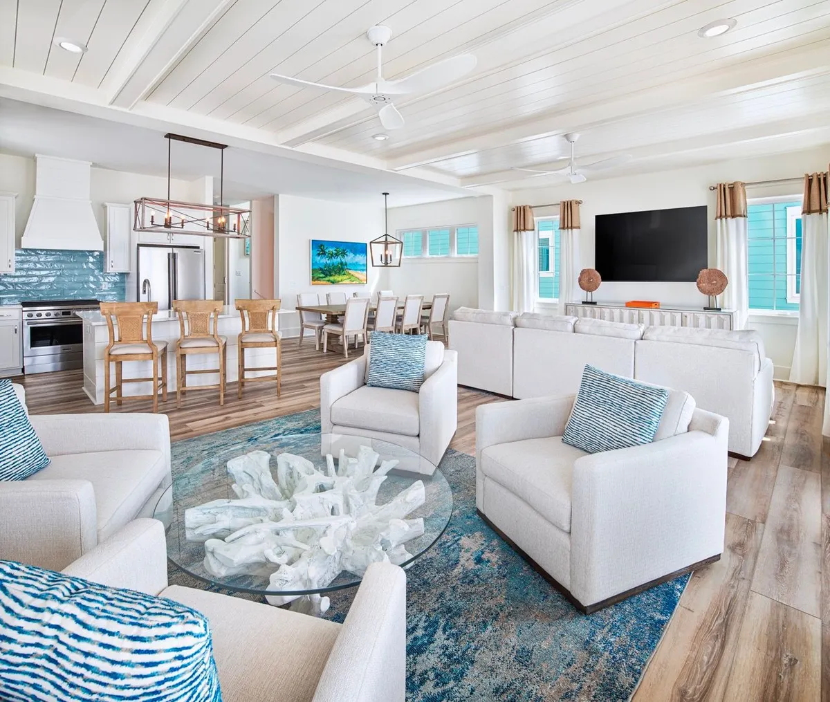 A bright, inviting open-concept living area featuring cushioned white armchairs with blue patterned pillows, a glass coffee table with a coral-inspired centerpiece, a large flat-screen TV on the wall, and a kitchen island with wooden bar stools. The space seamlessly connects the living room, kitchen, and dining area, with a fresh beachy vibe throughout.