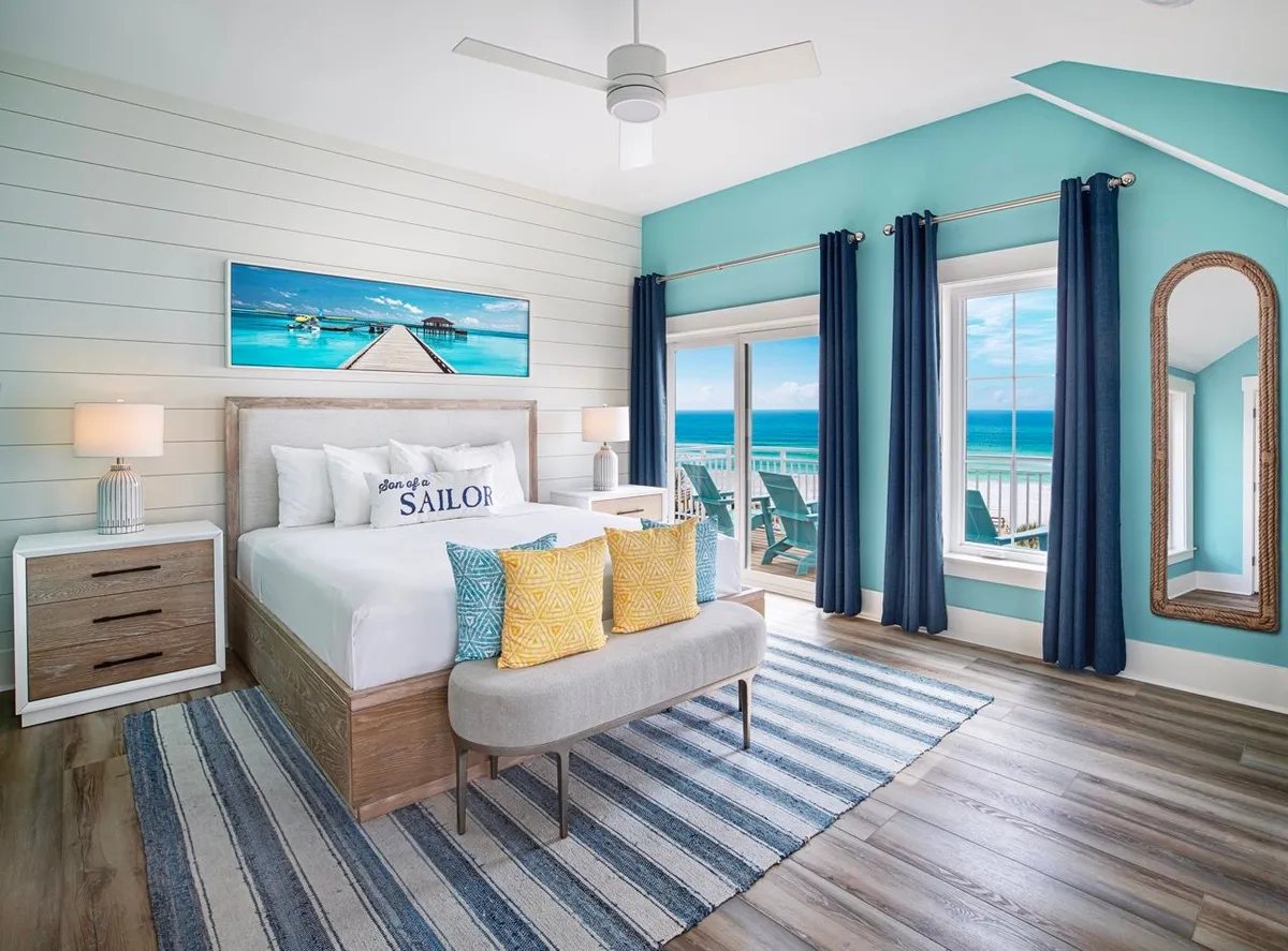 A spacious bedroom featuring a large comfortable bed with nautical-themed pillows, floor-to-ceiling windows that offer a stunning ocean view, and a private balcony accessible from the room. The room is decorated with a striped area rug and a natural wood-framed arched mirror, creating a relaxing and upscale coastal retreat.