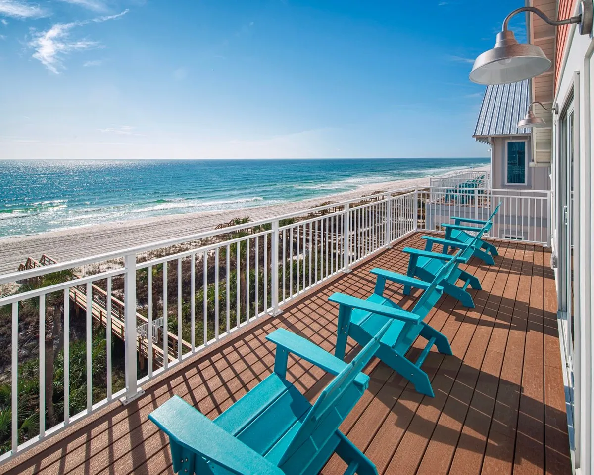 A serene beachfront balcony lined with blue Adirondack chairs, white railing, and a wooden deck floor. The view stretches out to an expansive ocean and sandy beach, inviting relaxation and outdoor enjoyment just steps from the main living area.