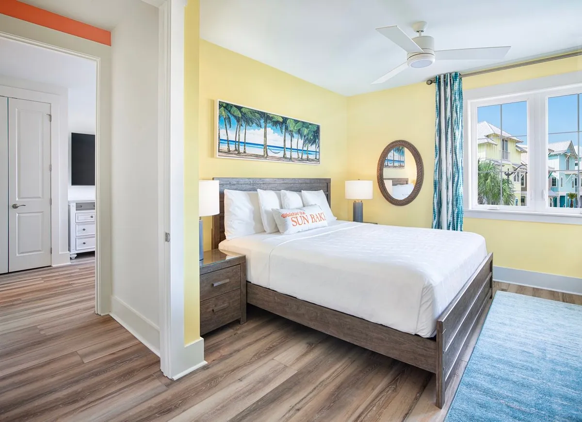 A king-size bed dressed in crisp white linens sits beneath beach-themed wall art, with a round mirror beside a large window that offers views of colorful beach houses outside. The space is airy and inviting, combining comfort with coastal charm.