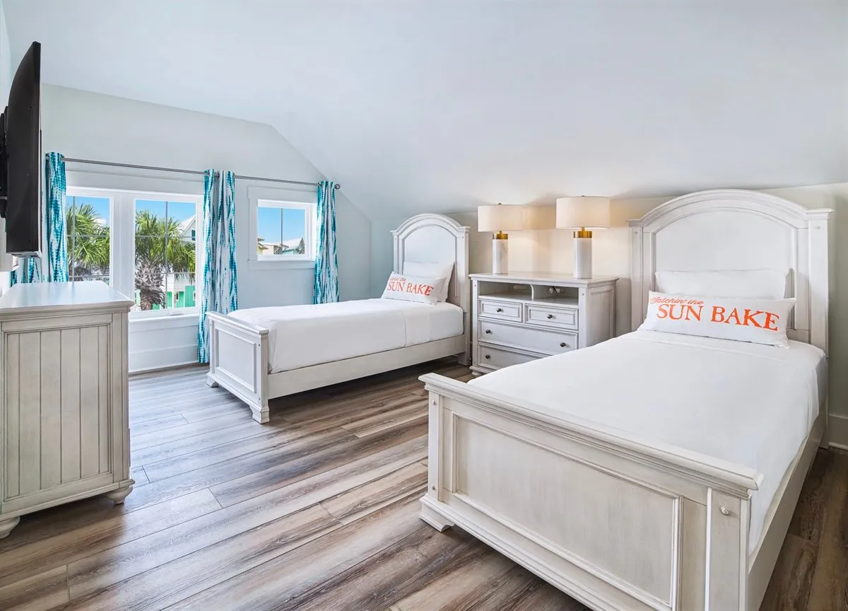 A serene twin bedroom with two white beds adorned with decorative pillows. Large windows let in natural light, enhanced by blue patterned curtains, and the space is styled with a white dresser, nightstand, and wood-style flooring for a clean, beach-themed look.