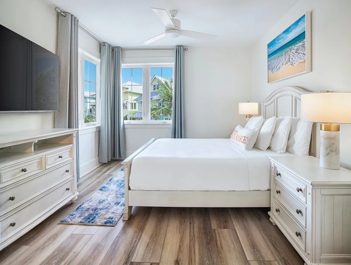 A spacious, sunlit villa bedroom featuring a king-sized bed with crisp white linens, large windows offering views of neighboring beach houses and palm trees, a wall-mounted flat-screen TV, and beach-themed artwork. The room exudes comfort, privacy, and coastal elegance.