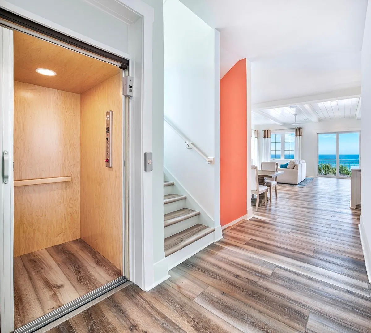 A bright, modern recreation room interior featuring an open elevator with wood paneling, a coral accent wall, and a glimpse of a living area with large windows displaying an ocean view. The space feels open, cheerful, and conducive to indoor fun and relaxation.