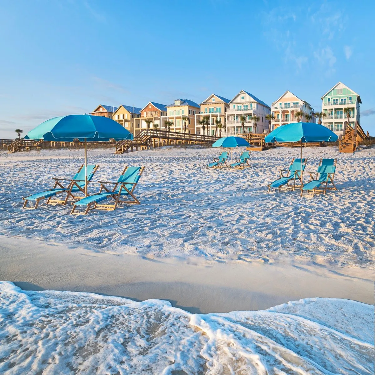 This image features a row of colorful beach cottages with balconies facing the ocean, bright blue umbrellas and lounge chairs neatly arranged on the soft white sand, and wooden dune walkovers connecting the cottages directly to the beach. Gentle waves roll onto the shore, and the setup invites guests to relax and enjoy the sun and surf right outside their accommodations.