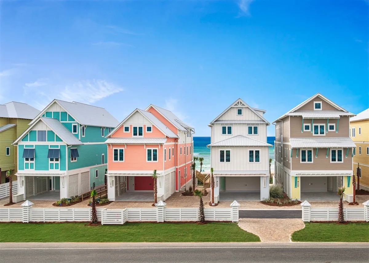 A row of colorful, modern three-story beach houses with private beach access, white picket fences, and lush landscaped yards. The inviting walkway leads towards the vibrant blue ocean and sky, suggesting easy access to nearby attractions and settings for group adventures.
